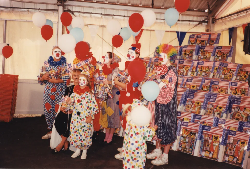 heritage-molly-browns-fireworks-commercial Historic photo of costumed clowns and children holding balloons and small American flags inside a Molly Brown’s Fireworks tent.