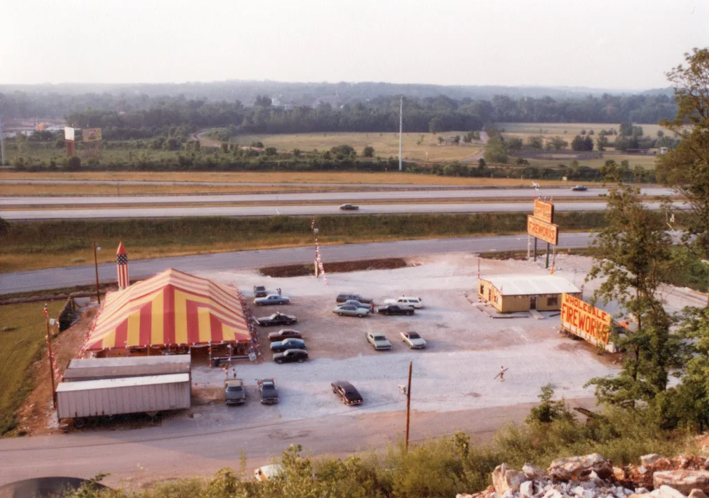 heritage-molly-browns-valley-park Historic aerial photo of a Molly Brown’s Fireworks location with a large striped tent, parking lot, roadside fireworks signs, and highway in the background.