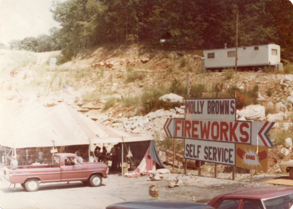 heritage-molly-browns-valley-park-2 Faded historic photo of a Molly Brown’s Fireworks self-service roadside tent with a large fireworks sign, a pink pickup truck, and a rocky hillside behind it.
