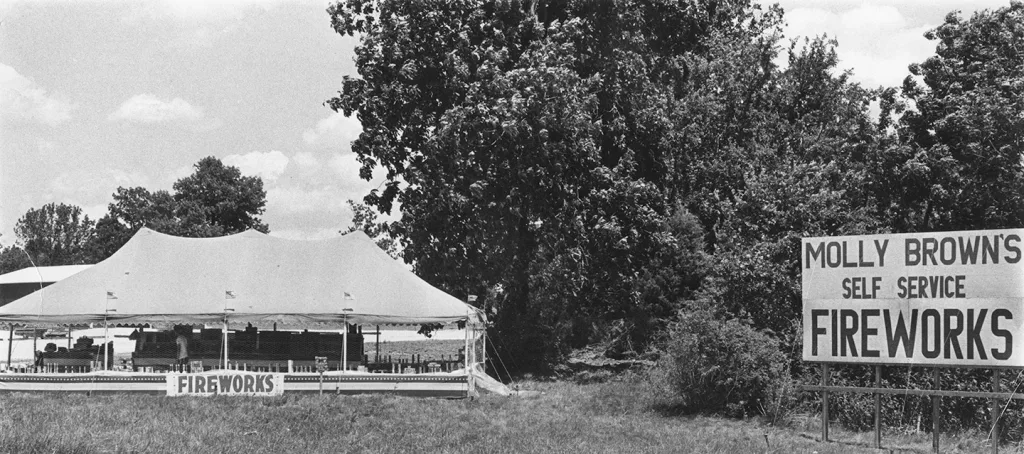 heritage-molly-browns-self-service-fireworks-2 Historic black-and-white photo of a Molly Brown’s Fireworks roadside tent with a large self-service fireworks sign beside a wooded area.