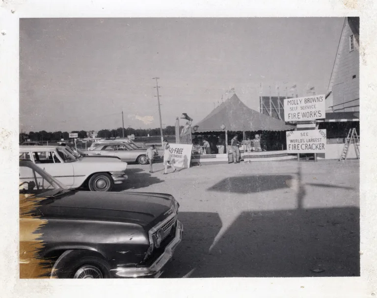 Historic black-and-white photo of a Molly Brown’s Fireworks roadside tent with vintage cars parked nearby and signs advertising self-service fireworks and the world’s largest firecracker.
