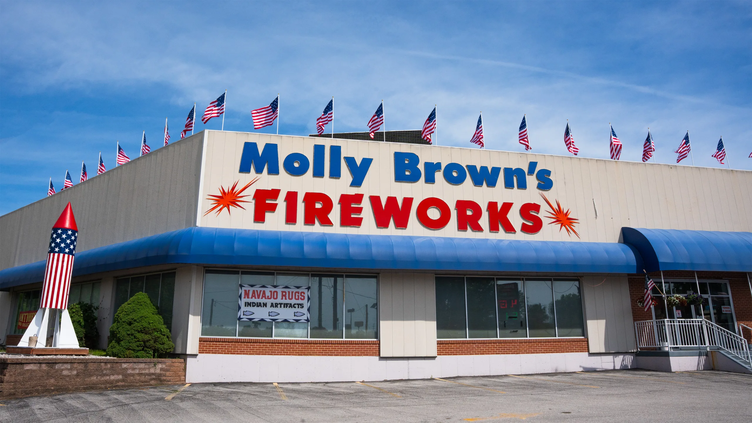 Exterior of the Molly Brown’s Fireworks store with large red and blue signage, a blue awning, and American flags flying along the roof.