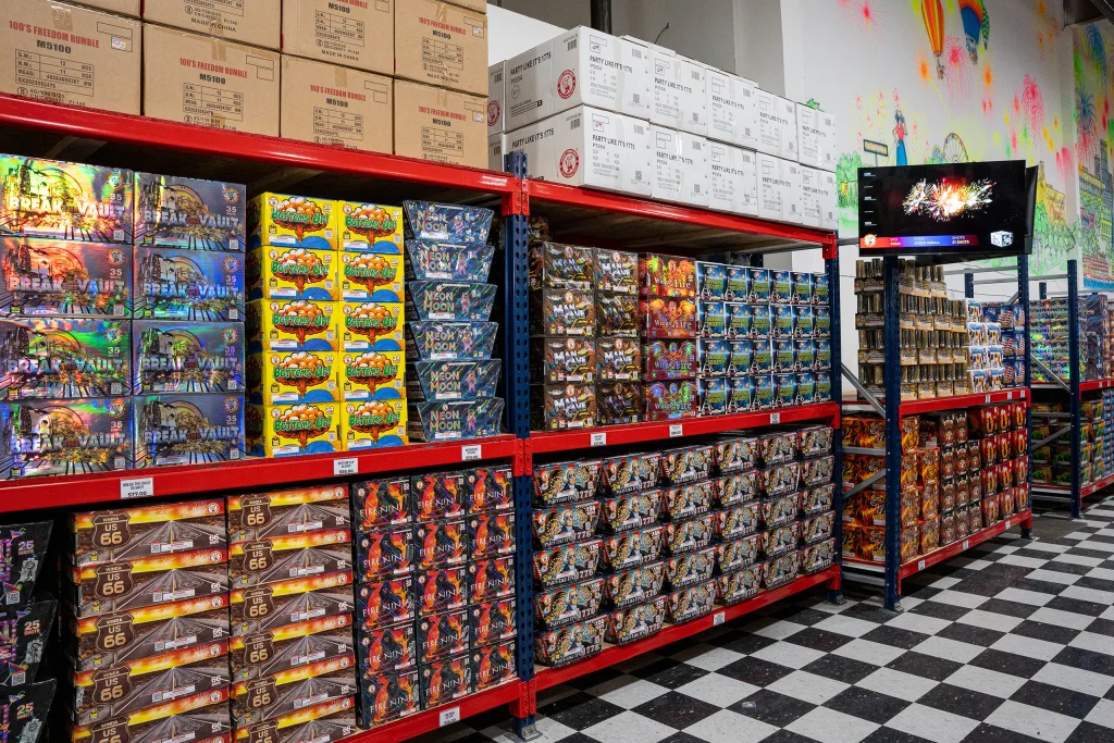 photo-molly-browns-fireworks-interior-1 Interior aisle at Molly Brown’s Fireworks showing shelves stocked with colorful boxed fireworks, bulk cases above, checkered flooring, and an in-store video display.