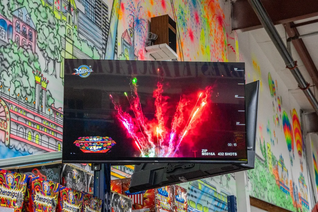 photo-molly-browns-fireworks-interior-3 In-store video display at Molly Brown’s Fireworks showing a fireworks product demo, mounted above boxed fireworks with a colorful mural in the background.