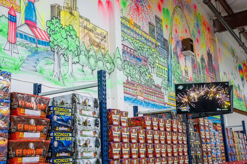 photo-molly-browns-fireworks-interior-4 Molly Brown’s Fireworks interior wall display with stacked boxed fireworks, an in-store video demo screen, and a large colorful mural of St. Louis landmarks and fireworks.