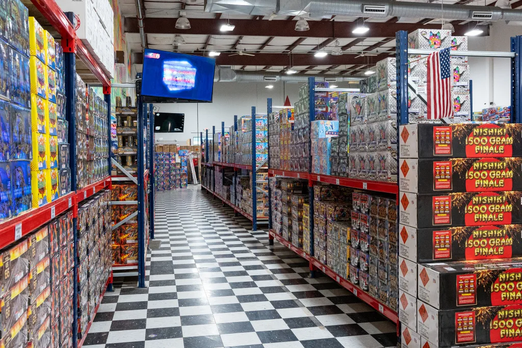 photo-molly-browns-fireworks-interior-5 Wide aisle inside Molly Brown’s Fireworks showing tall shelves stocked with fireworks, black-and-white checkered flooring, and video display screens.